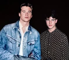 two young men standing next to each other in front of a black background and one is wearing a denim jacket
