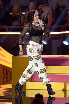 a woman in white pants and black top holding a microphone on stage with other people behind her