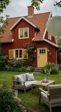 a red house with two chairs and a table in front of it, surrounded by greenery