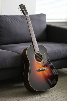 an acoustic guitar sitting on the floor in front of a couch with a window behind it