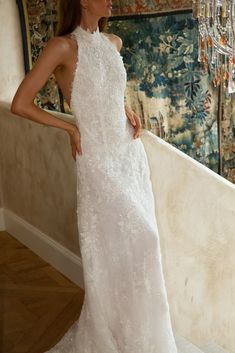 A woman wearing an elegant, lace halter-neck wedding gown stands by a staircase. A floral tapestry and a crystal chandelier are in the background.