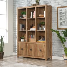 a wooden bookcase with many books and plants on it in front of a brick wall