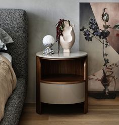 a white vase sitting on top of a wooden table next to a painting and a bed