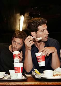 two men sitting at a table eating food and drinking from cups with napkins in front of them