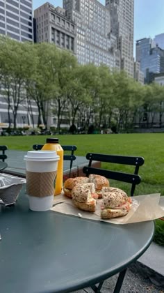 sandwiches and drinks are sitting on a table in the middle of a park with skyscrapers in the background