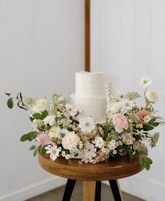a white wedding cake with flowers and greenery on a wooden stand in front of a white wall