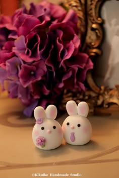 two little white rabbits sitting next to each other on a table with purple flowers in the background