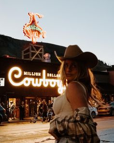 a woman wearing a cowboy hat standing in front of a building with neon signs on it