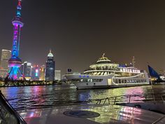 a boat traveling down the river in front of a large city at night with colorful lights on it