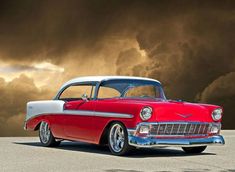 an old red and white car is parked in front of a dark cloud filled sky