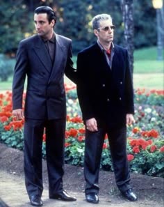 two men standing next to each other in front of red flowers and green grass with trees behind them