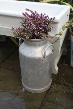 a metal watering can with purple flowers in it next to a white bench and potted plant
