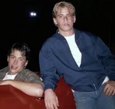 two young men sitting on top of a red car in front of a dark background