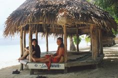 two people sitting on a bench under a straw hut