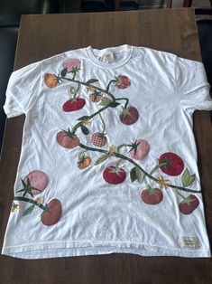 a white t - shirt with strawberries and cherries embroidered on the front, sitting on a wooden table