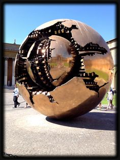 a large metal ball sitting in the middle of a courtyard with people walking around it