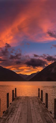 a wooden dock sitting on top of a lake under a colorful sky with clouds in the background