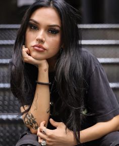 a woman with long black hair and tattoos on her arm, sitting in front of stairs