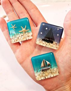 three small glass magnets in the palm of someone's hand with a sailboat and starfish on them