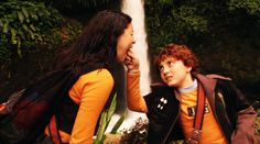 two people standing next to each other in front of a waterfall