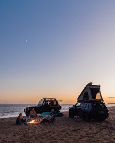 two people are sitting on the beach next to their camper van at sunset with an open hatchback