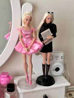 two barbie dolls standing on top of a white dresser next to a mirror and camera
