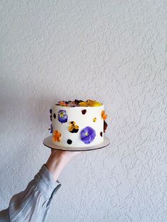 a person holding a cake with white frosting and purple flowers on it in front of a wall