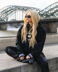 a blonde haired woman sitting on the edge of a wall next to a river and bridge