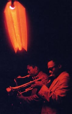 two men playing instruments in front of a bright orange light on a black background with an object hanging from it's side