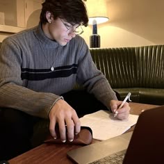 a young man sitting at a table with a laptop and pen in his hand while writing on paper