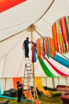 a man standing on a ladder in front of a tent with colorful streamers hanging from it