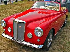 an old red convertible car parked on top of a grass covered field next to people