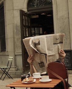 a person sitting at a table reading a newspaper and holding a coffee cup in front of them