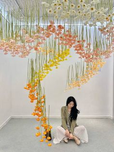 a woman sitting on the ground in front of a suspended flower sculpture with flowers hanging from it's ceiling