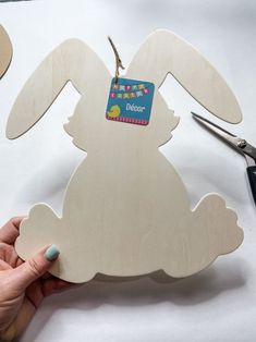 a hand holding a wooden cutout of a bunny with scissors next to it on a table