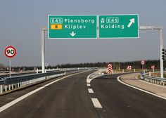 two green highway signs on the side of an empty road with trees in the background