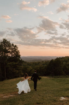 a bride and groom walking in the grass at sunset