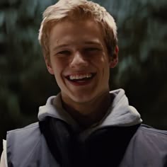 a young boy with blonde hair smiling and wearing a black vest in front of trees