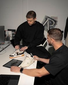 two men sitting at a table with shoes on it and an open laptop in front of them