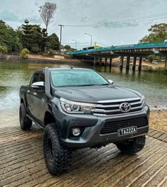 a grey toyota hilux is parked in front of a body of water with a bridge in the background