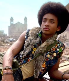a young man with an afro is sitting on the ground in front of destroyed buildings