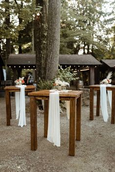 two wooden tables with white sashes are set up for an outdoor wedding reception in the woods