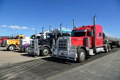several semi trucks are lined up in a row on the pavement, one is red and yellow