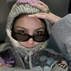 a woman wearing sunglasses and a knitted hoodie holds her hands up to her head