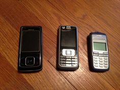 three old cell phones sitting on top of a wooden table next to each other, one black and one silver