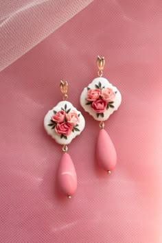 two pink and white earrings with roses on them sitting on a pink cloth covered surface