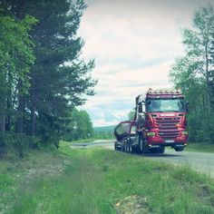 a red semi truck driving down a rural road next to tall grass and trees on either side of the road