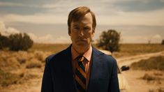 a man wearing a suit and tie standing in the middle of a dirt road next to a car