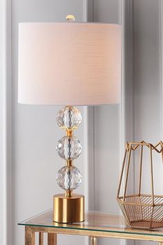 a glass table with a lamp on it and a basket next to it in front of a white wall