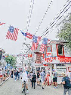 many people are walking and riding bikes down the street with american flags hanging above them
